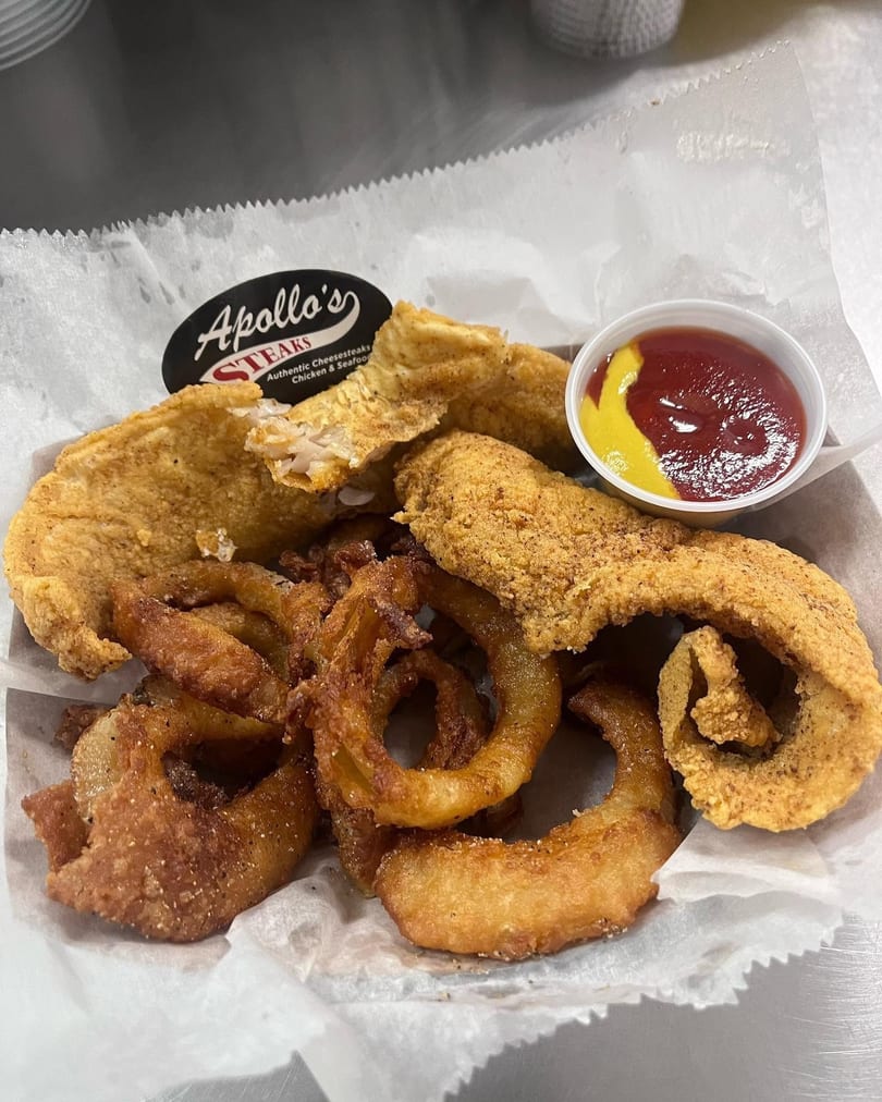 Apollo's Steaks fried fish and onion rings with ketchup and mustard in a white paper-lined basket.