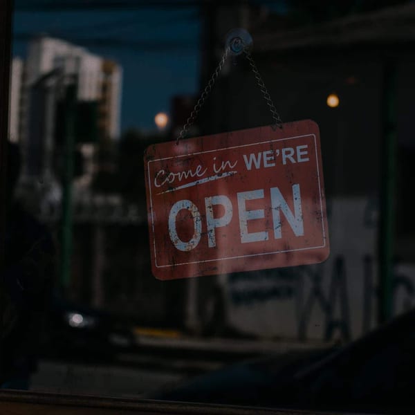 A weathered red "Come in We're OPEN" sign hangs on a glass door, reflecting a blurry street.